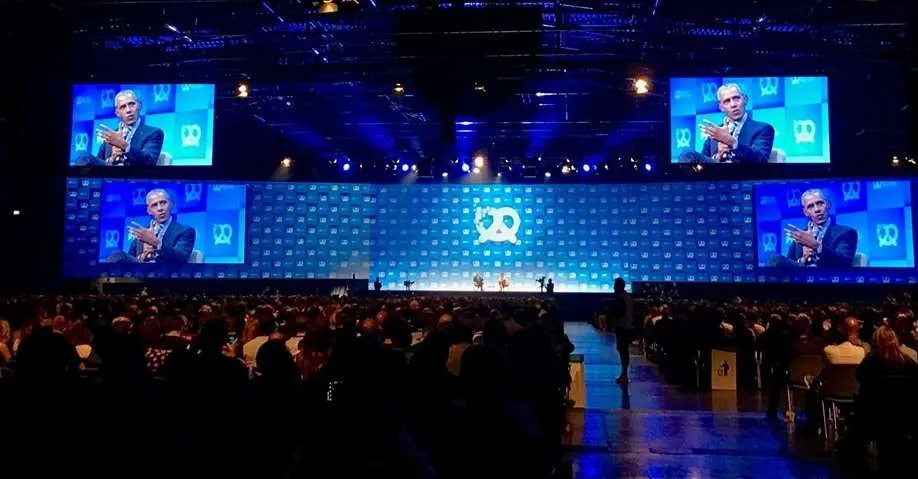 Barack Obama speaking on the Bits & Pretzels stage, shown on the big screens above a packed hall.
