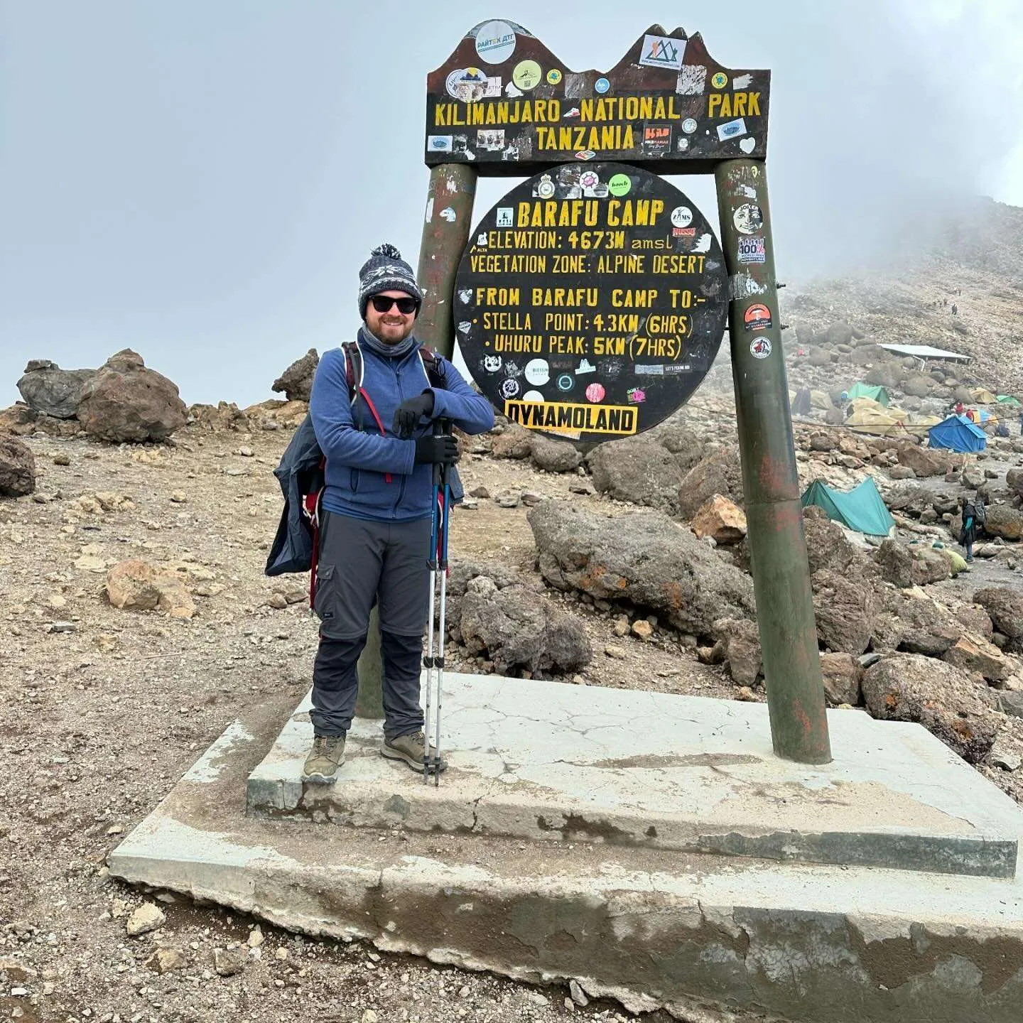 A climber in a blue jacket and hat beside the Barafu Camp sign in Kilimanjaro National Park, on rocky ground in the mist.