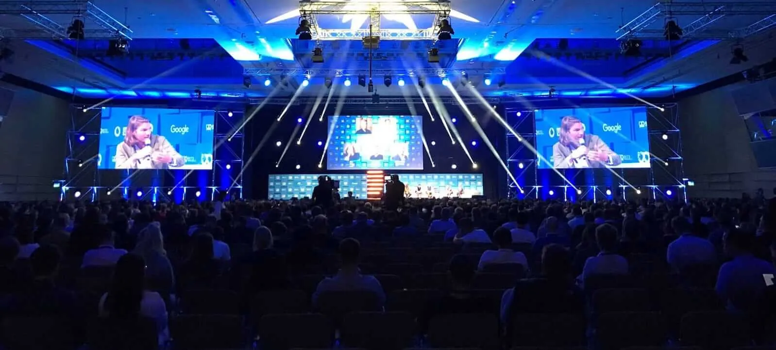 A packed Bits & Pretzels hall facing a stage lit in blue, a panel speaker shown on the big screens either side.