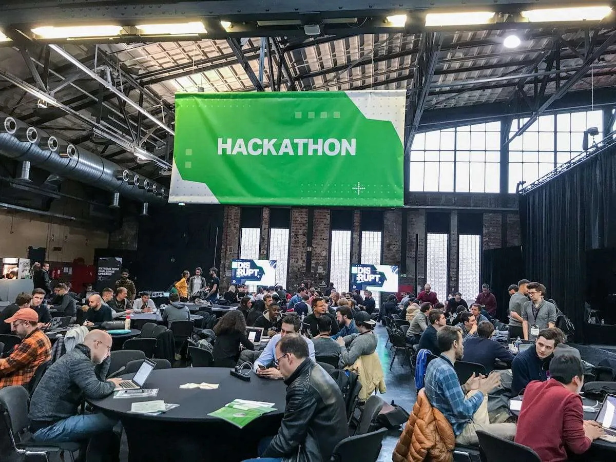 Developers at round tables with laptops in the Disrupt Berlin hackathon hall, under a green banner reading “Hackathon”.
