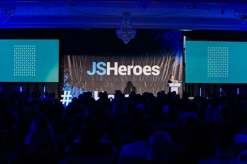 A speaker at a lectern on the JSHeroes stage in Cluj, under chandeliers and between two screens of binary digits, before a darkened audience.