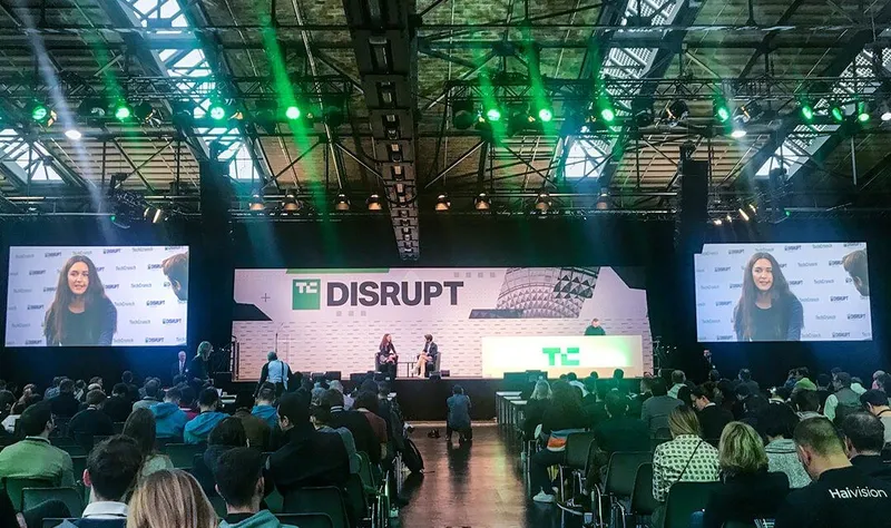 The Disrupt Berlin main stage under green lights and the TechCrunch “Disrupt” sign, a speaker on the big screens either side of a seated audience.