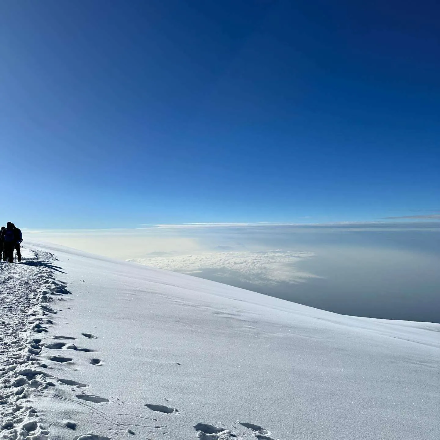 Climbers walking along a snowy ridge high on the mountain, above a sea of clouds, under a clear blue sky.