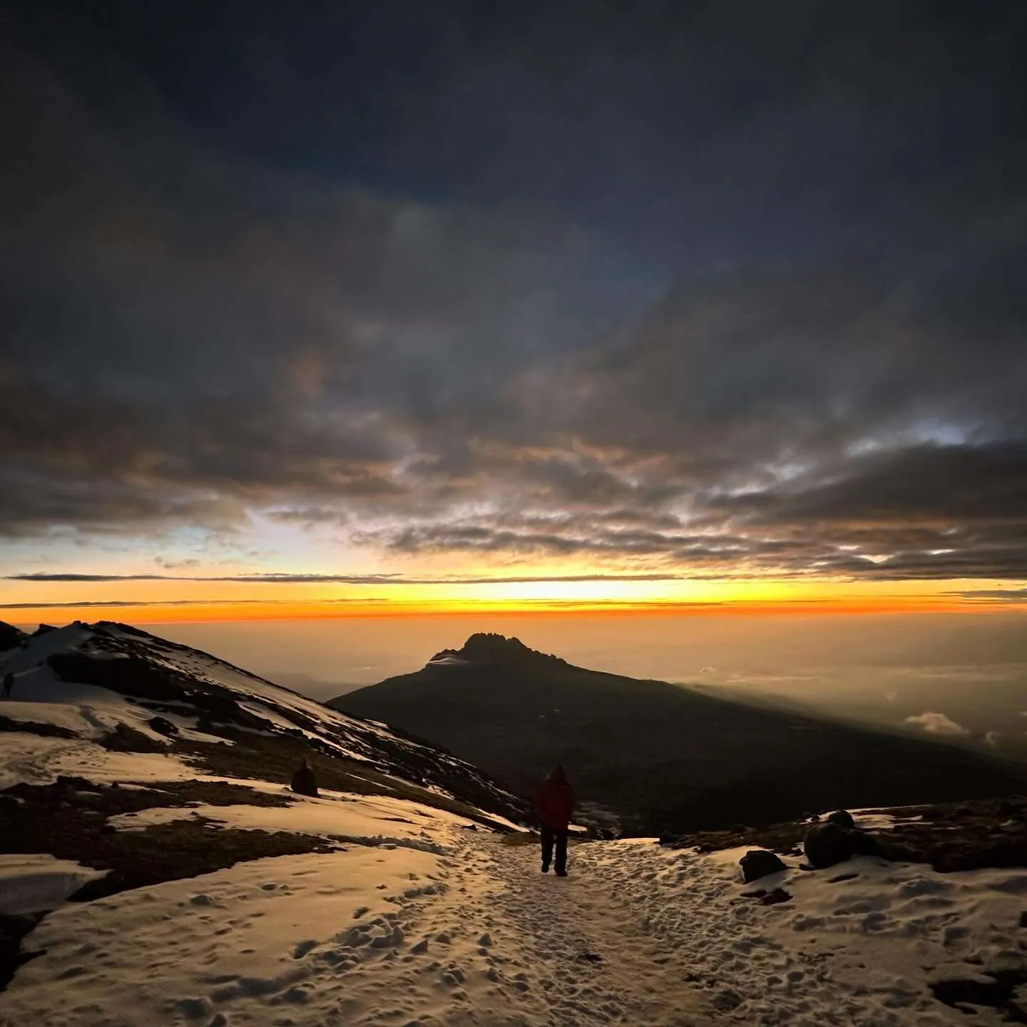 A lone climber on the snowy trail at dawn, with a mountain peak ahead and a band of orange light under dark clouds.