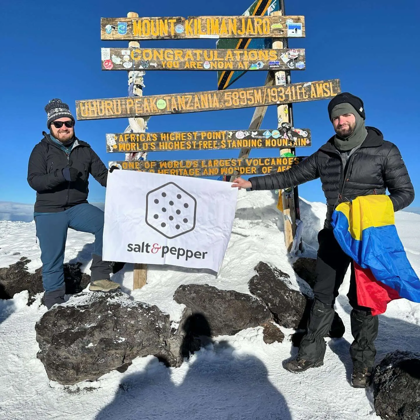 Two climbers at the Uhuru Peak summit sign holding up a Salt &amp; Pepper flag, one of them wrapped in the Romanian flag.