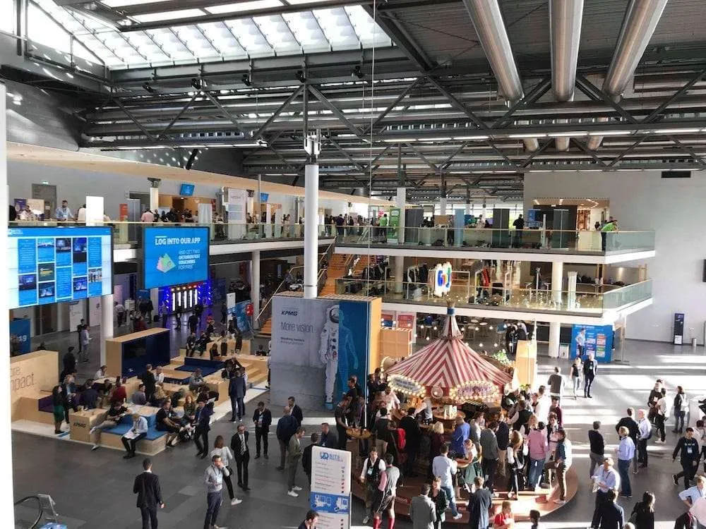 The Bits & Pretzels venue in Munich seen from a balcony, attendees mingling around lounges and a red-and-white striped carousel bar.